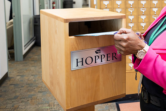 Hand placing bill documents in to a wooden box labeled 'hopper'